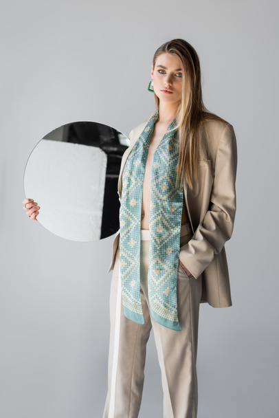 jeune femme en costume tenant miroir rond et debout avec la main dans la poche sur gris - Photo, image