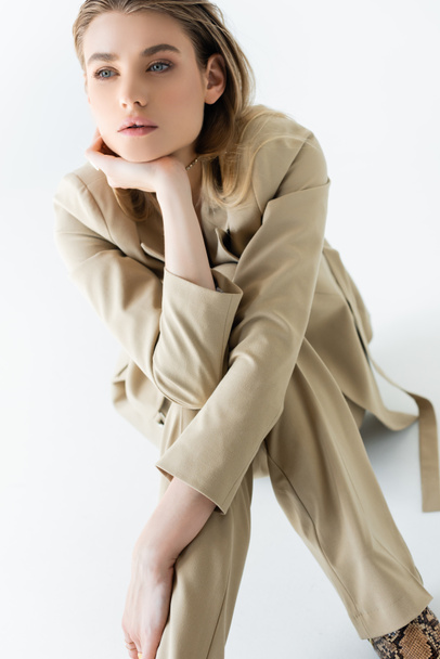 stylish and dreamy model in beige suit sitting on white - Foto, Bild