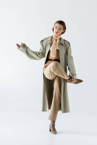 full length of young woman in glasses, trench coat and scarf posing on white - Foto, afbeelding