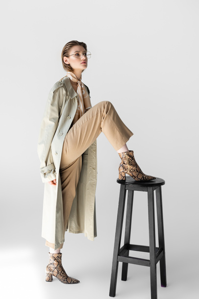 trendy woman in trench coat and scarf posing near chair on white - Fotó, kép