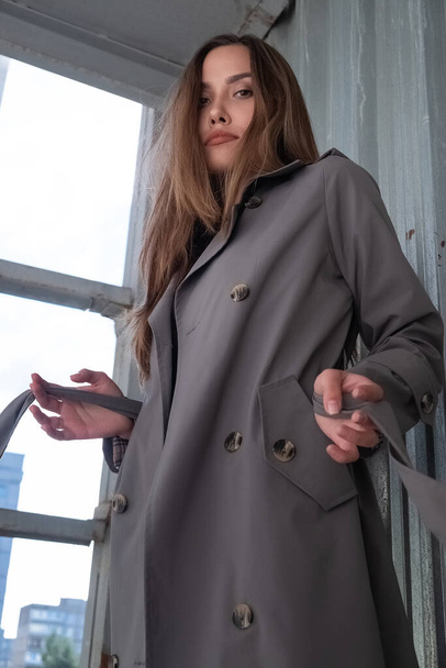 приваблива молода азіатська модель в сірому траншеї пальто, що стоїть біля стіни на відкритому повітрі
 - Фото, зображення