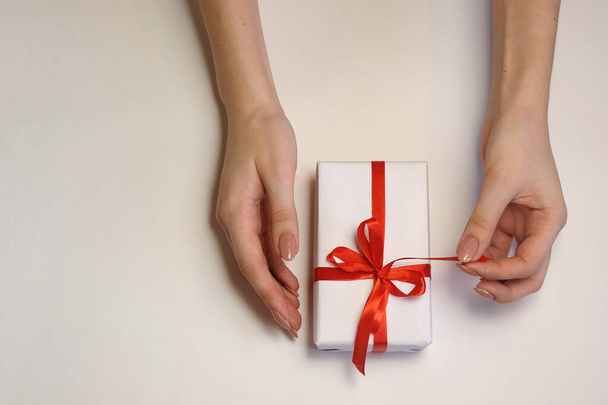 vrouwelijke handen maken het rode lint los op een geschenkdoos op een lichte achtergrond. Kerstmis en Nieuwjaar of seizoen groeten. Selectieve focus - Foto, afbeelding