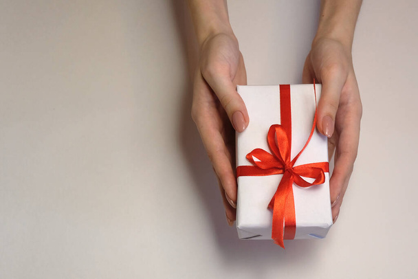 manos femeninas con una caja de regalo y una cinta roja sobre un fondo claro. Navidad y Año Nuevo o saludo de temporada. Enfoque selectivo - Foto, Imagen