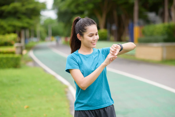 A happy young woman in sportswear using smart watch while exercising in park - Valokuva, kuva