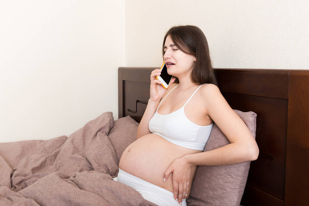 Emotionaalinen raskaana oleva nainen itkee ja puhuu matkapuhelimella kotona sängyssä. - Valokuva, kuva