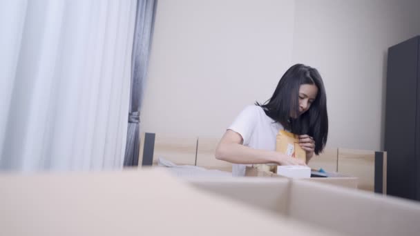 Asian young woman sit down on comfortable bed moving inside new place with parcel box on her lap, taking things out of the box, new apartment, Sorting cleaning, Relocating moving, stay at home - Footage, Video