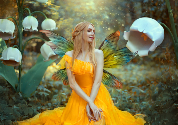 Portrait of happy fantasy woman blonde forest fairy. Fashion model in a bright yellow dress with butterfly wings sits posing in nature. Large flowers scenery decor white lilies. Light magic radiance. - Valokuva, kuva