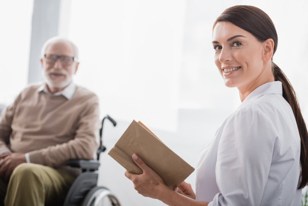 usmívající se sociální pracovník při pohledu na kameru při čtení knihy na staré zdravotně postižené muže na rozmazaném pozadí - Fotografie, Obrázek