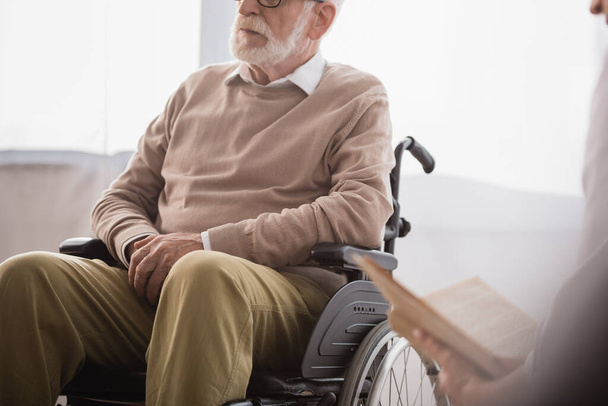 bijgesneden beeld van sociaal werker leesboek voor gehandicapte man in rolstoel op wazige voorgrond - Foto, afbeelding