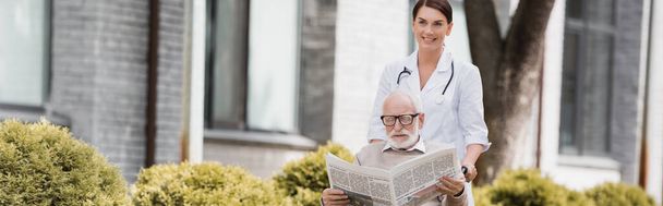 Neşeli yaşlı hemşire engelli bir adamla yürüyor gazete ve afiş okuyor. - Fotoğraf, Görsel