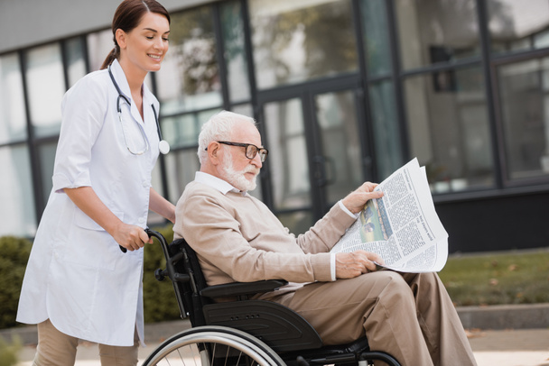 handicapped aged man reading book in wheelchair while walking with geriatric nurse outdoors - Photo, Image