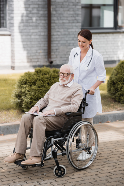 ruskeaverikkö sosiaalityöntekijä kävelee ikääntyneen vammaisen miehen kanssa pyörätuolissa ulkona - Valokuva, kuva