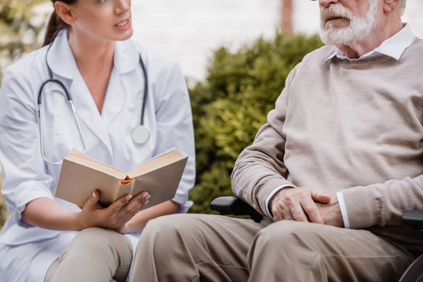 cropped view of geriatric nurse reading book to elderly man outside - Photo, Image