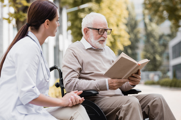 disabled man in eyeglasses reading book near social worker outdoors - Fotó, kép