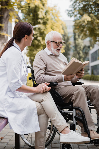 Altenpfleger sitzt neben älterem behinderten Mann und liest Buch im Freien - Foto, Bild