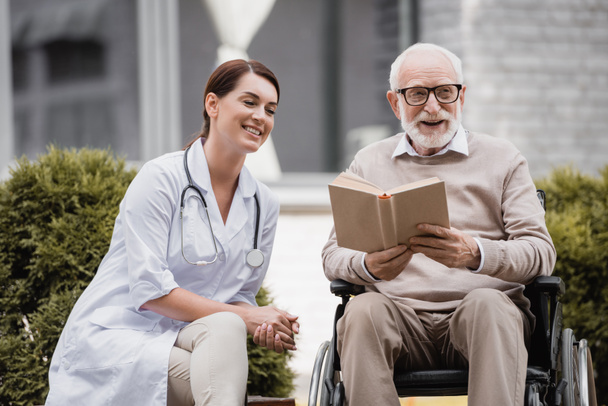 fröhliche Altenpflegerin nahe älterer behinderter Mann liest Buch im Rollstuhl im Freien - Foto, Bild