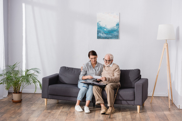 Älterer Vater mit erwachsener Tochter blättert im Fotoalbum, während er zu Hause auf dem Sofa sitzt - Foto, Bild