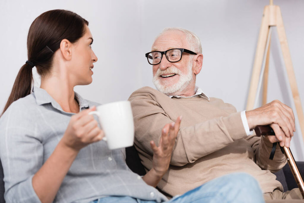 brunette woman holding tea and gesturing while talking to cheerful elderly father - Фото, изображение