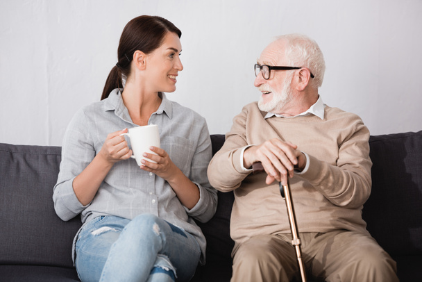 brunette woman with cup of tea and smiling aged man with walking stick talking while sitting on sofa at home - 写真・画像