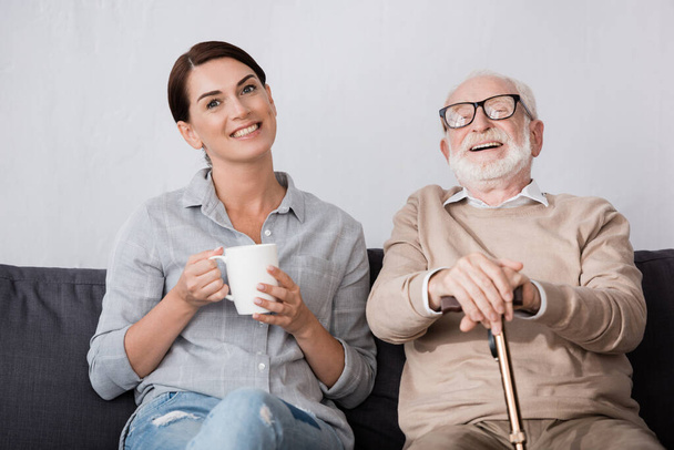 vrolijk vrouw en lachen bejaarde vader kijken naar camera terwijl zitten op de bank - Foto, afbeelding
