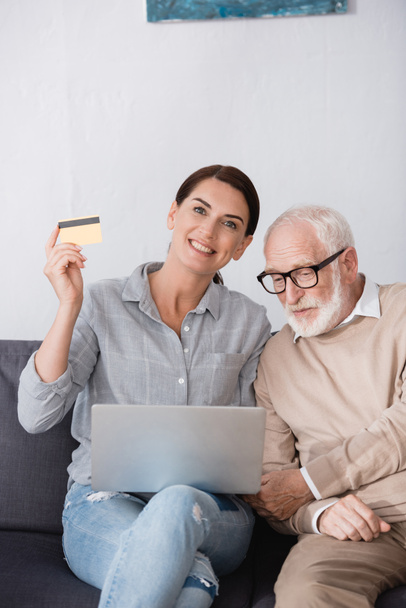 cheerful woman holding laptop and credit card while sitting near elderly father - Photo, Image