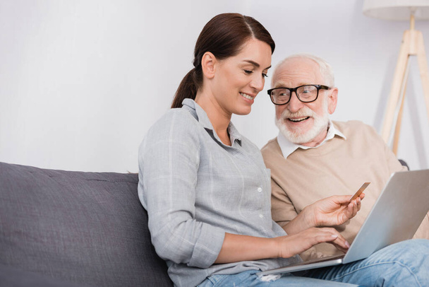 smiling woman using laptop and holding credit card near aged father at home - Foto, Imagem