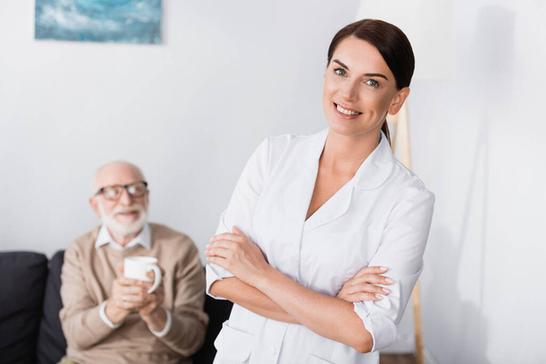 fröhliche Sozialarbeiterin lächelt in die Kamera, während sie mit verschränkten Armen neben einem alten Mann steht, der eine Tasse Tee hält - Foto, Bild