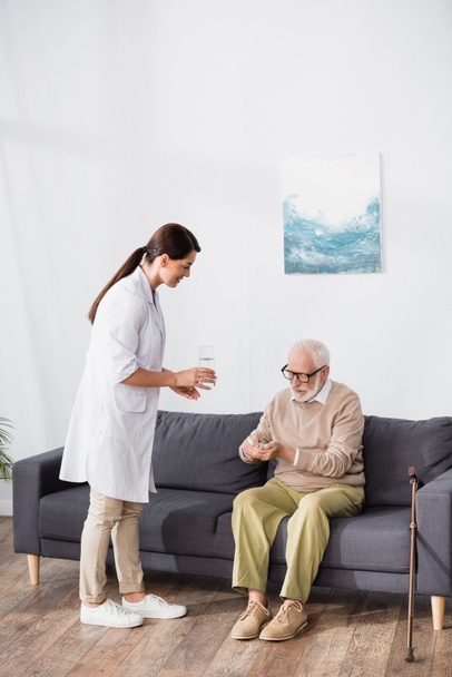 anciano sentado en el sofá y sosteniendo pastillas mientras asistente médico vaso de agua - Foto, Imagen