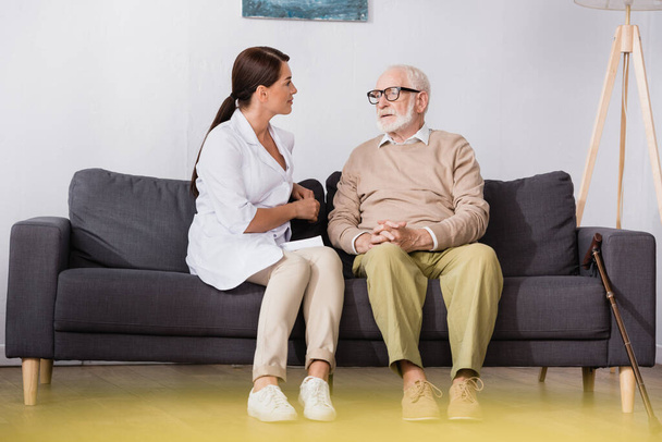 geriatric nurse talking to senior man while sitting on sofa at home on blurred foreground - Photo, Image