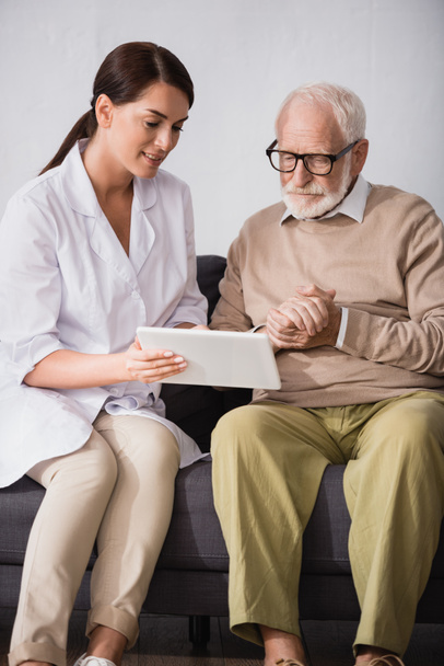 brunette geriatric nurse showing digital tablet to elderly man sitting with clenched hands - Foto, Bild