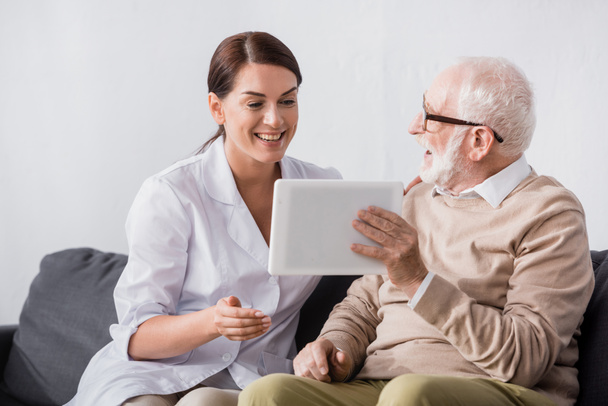 happy aged man showing digital tablet to excited social worker - Photo, Image