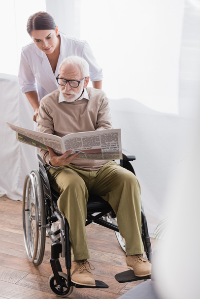 anciano discapacitado hombre leyendo periódico en silla de ruedas cerca de trabajador social - Foto, Imagen