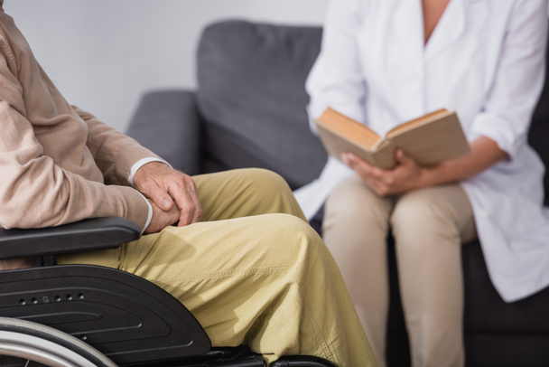 cropped view of geriatric nurse reading book to elderly man at home - Foto, imagen