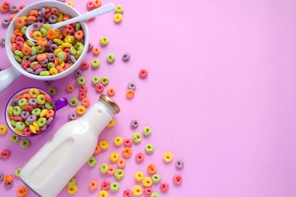 Una taza grande blanca y una ms pequea de color lila, ambas llenas de aros de cereal. Cerca a ellas una botella de leche fresca. - Foto, immagini