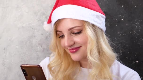 Gelukkig mooi blondje in santas hoed met behulp van haar smartphone, videogesprek, kijken naar het scherm en lachen. vakantie concept. close-up. - Video