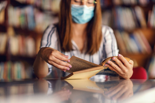 Young dedicated attractive brunette with face mask on sitting in library and reading a book. Alternative studying during corona pandemic concept. - Photo, Image