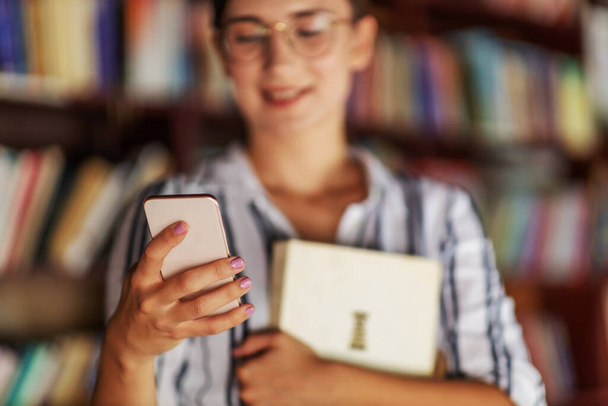 Kütüphanede elinde bir kitapla duran ve akıllı telefon kullanan çekici genç bir kızın yakın çekimi.. - Fotoğraf, Görsel