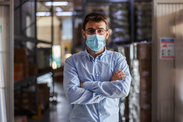 Young successful attractive businessman with surgical mask on standing in warehouse with arms crossed and looking at camera. Corona outbreak concept. - Φωτογραφία, εικόνα