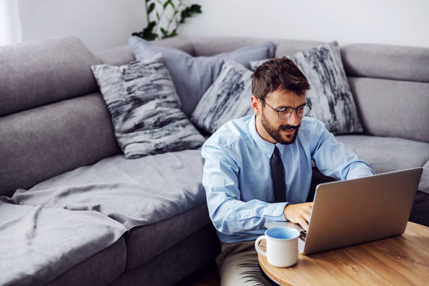 młody atrakcyjny brodaty biznesmen ubrany elegancko siedzi w domu na podłodze i pisze raport na laptopie. - Zdjęcie, obraz