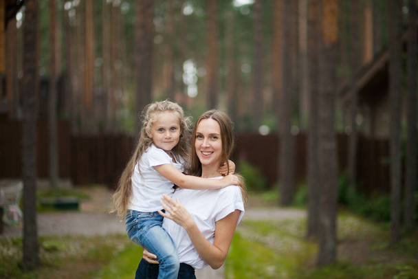 Happy smiling mother and her cute emotional little daughter child in white shirts and denim jeans are hugging and having fun outdoor in nature at countryside. Spending summer time with family - Photo, Image