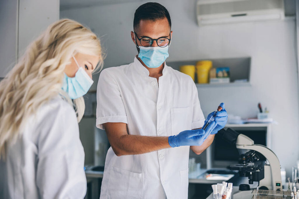 Dedykowany doświadczony męski asystent laboratoryjny z maską na twarzy i gumowymi rękawiczkami stojący w laboratorium i trzymający probówkę. Koncepcja epidemii wirusa korony. - Zdjęcie, obraz
