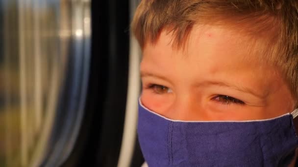電車に乗っている間に窓を見ている少年。綿のマスクの子供のクローズアップ。乗客の顔に太陽の光。輸送中の保護マスクを身に着けている子供.小さな旅行者の顔の感情 - 映像、動画