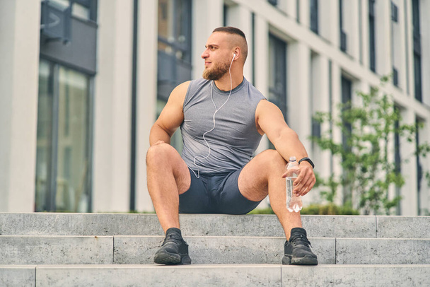 A guy in a sports uniform drinks water after a run while sitting on the stairs near a high-rise building. - Photo, image