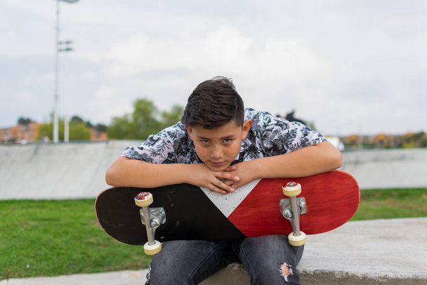 Уличное фото молодого красивого латиноамериканца с потрясающей стрижкой, держащего в руках синий красный белый скейтборд в листовой рубашке, черные порванные джинсы, черные туфли на коньках в скейт-парке Богота, Колумбия - Фото, изображение