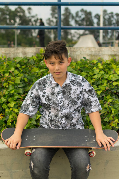 Street photo of young handsome hispanic asian boy with awesome haircut holding blue red white skateboard wearing leaf shirt, black ripped jeans, black skating shoes at skate park Bogota, Colombia - Foto, Imagen