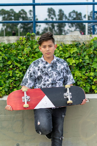 Straßenfoto von jungen hübschen hispanischen asiatischen Jungen mit genial Frisur hält blau rot weißes Skateboard tragen Blatthemd, schwarze zerrissene Jeans, schwarze Eislaufschuhe im Skatepark Bogota, Kolumbien - Foto, Bild