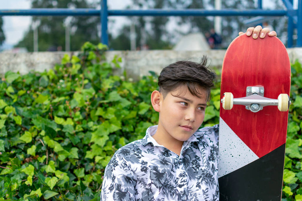 Street photo of young handsome hispanic asian boy with awesome haircut holding blue red white skateboard wearing leaf shirt, black ripped jeans, chaussures de patinage noires au skate park Bogota, Colombie - Photo, image