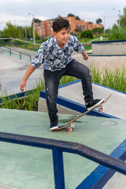 Street photo of young handsome hispanic asian boy with awesome haircut holding blue red white skateboard wearing leaf shirt, black ripped jeans, black skating shoes at skate park Bogota, Colombia - Foto, imagen