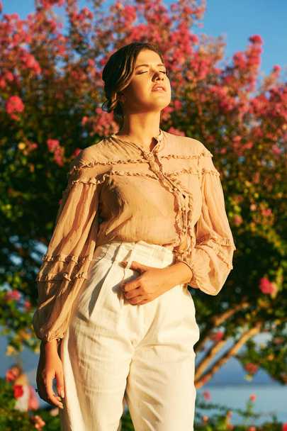 Outdoor mode portret van stijlvolle jonge vrouw dragen beige blouse en witte broek, poseren tegen roze bloeiende boom, zonsondergang licht - Foto, afbeelding