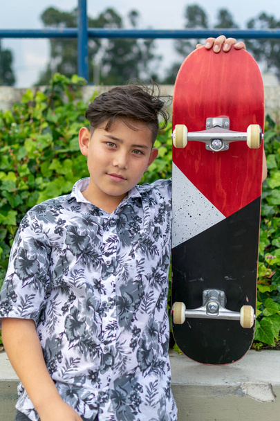 Street photo of young handsome hispanic asian boy with awesome haircut holding blue red white skateboard wearing leaf shirt, black ripped jeans, black skating shoes at skate park Bogota, Colombia - Photo, Image